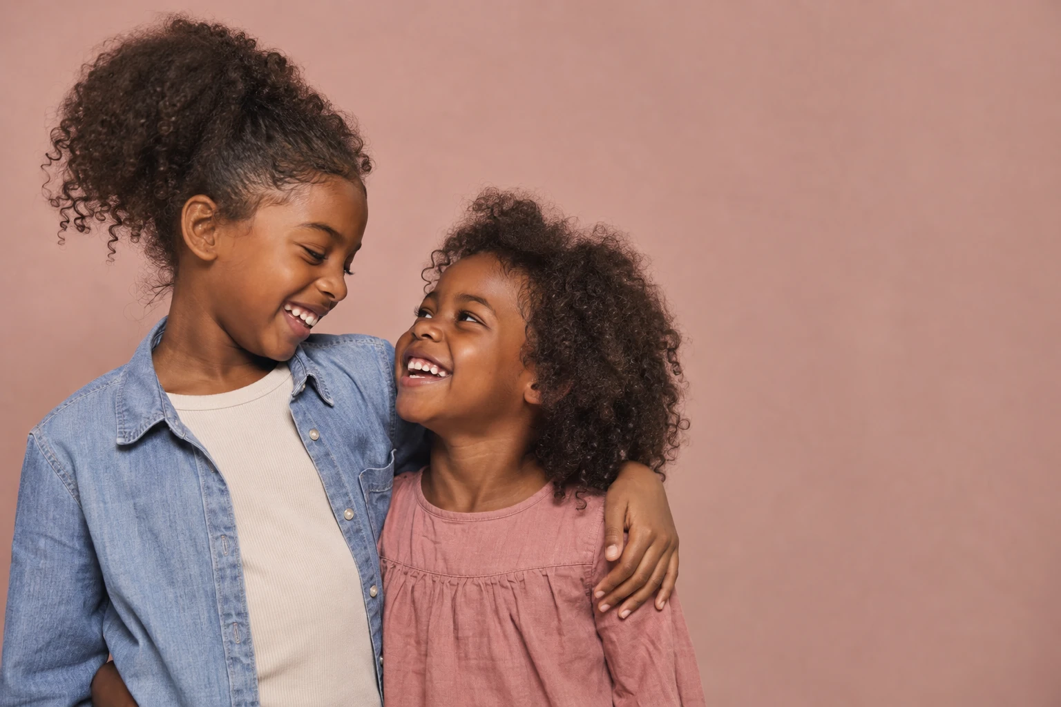Two smiling girls with their arms around one another against a pink background.