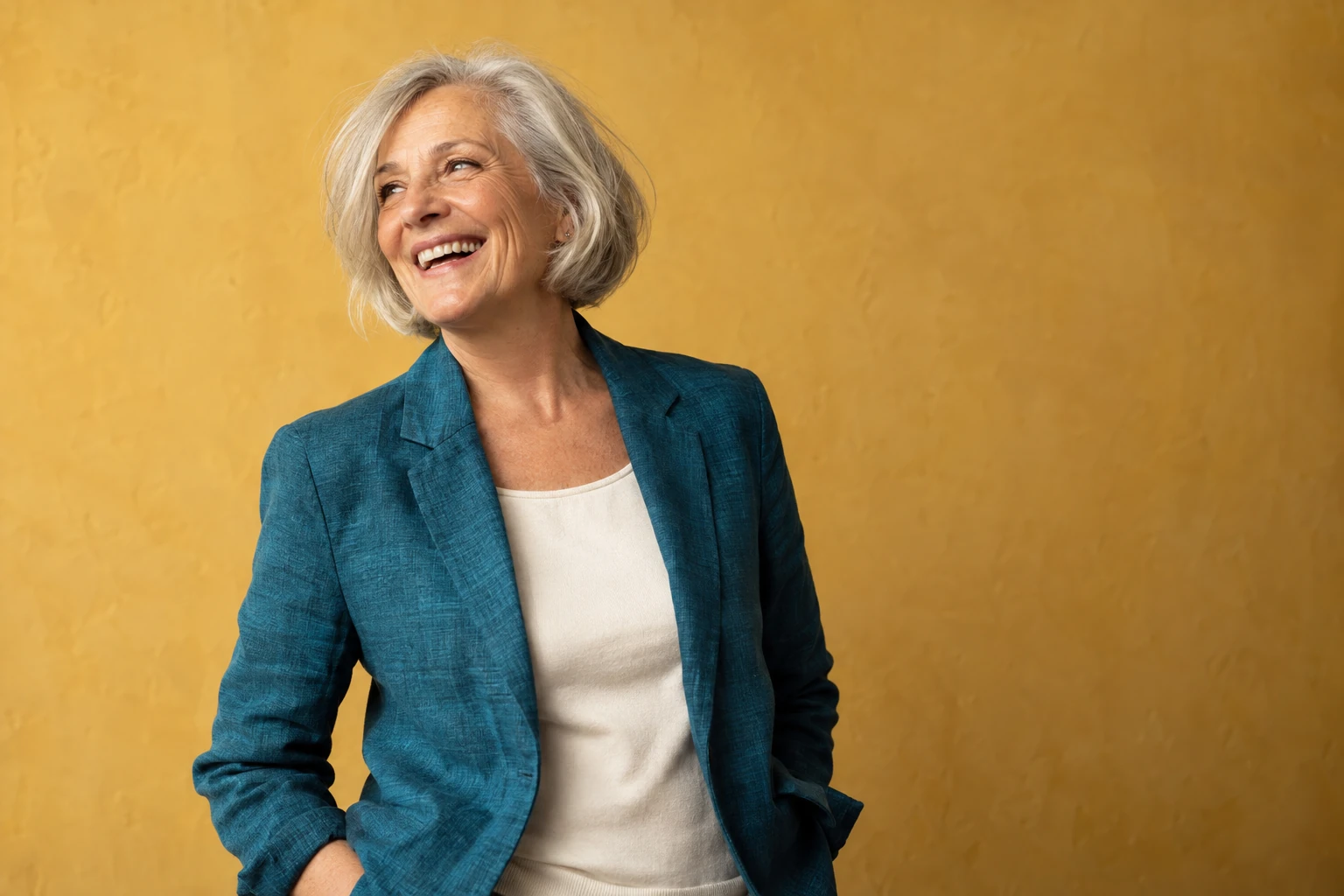 Smiling older woman in a teal jacket against a warm yellow background.